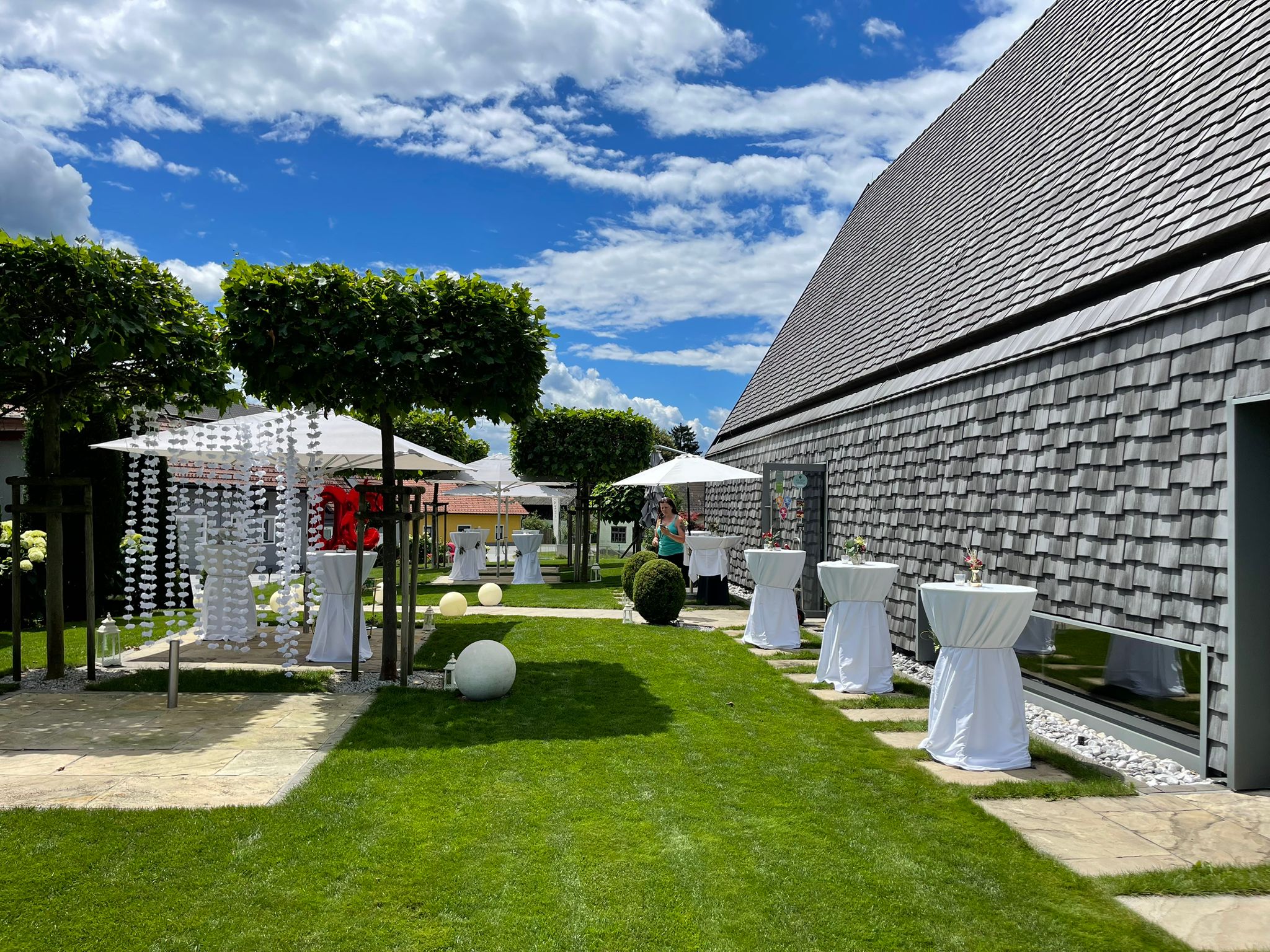 Garten mit dekorierten Stehtischen, weißen Kugelleuchten und moderner Buschenschank-Fassade.